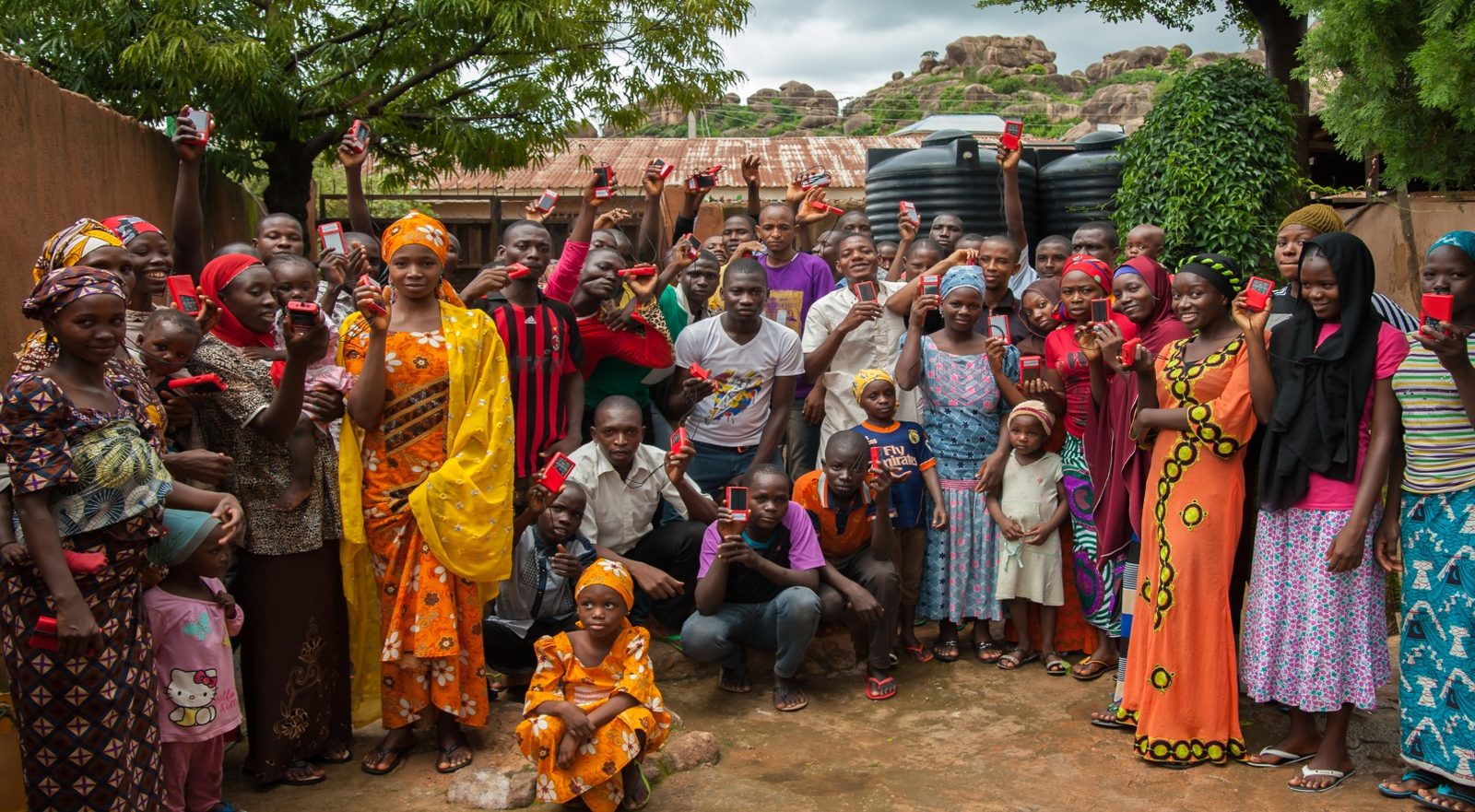 A village community gathered outdoors, joyfully holding up their red Galcom audio players